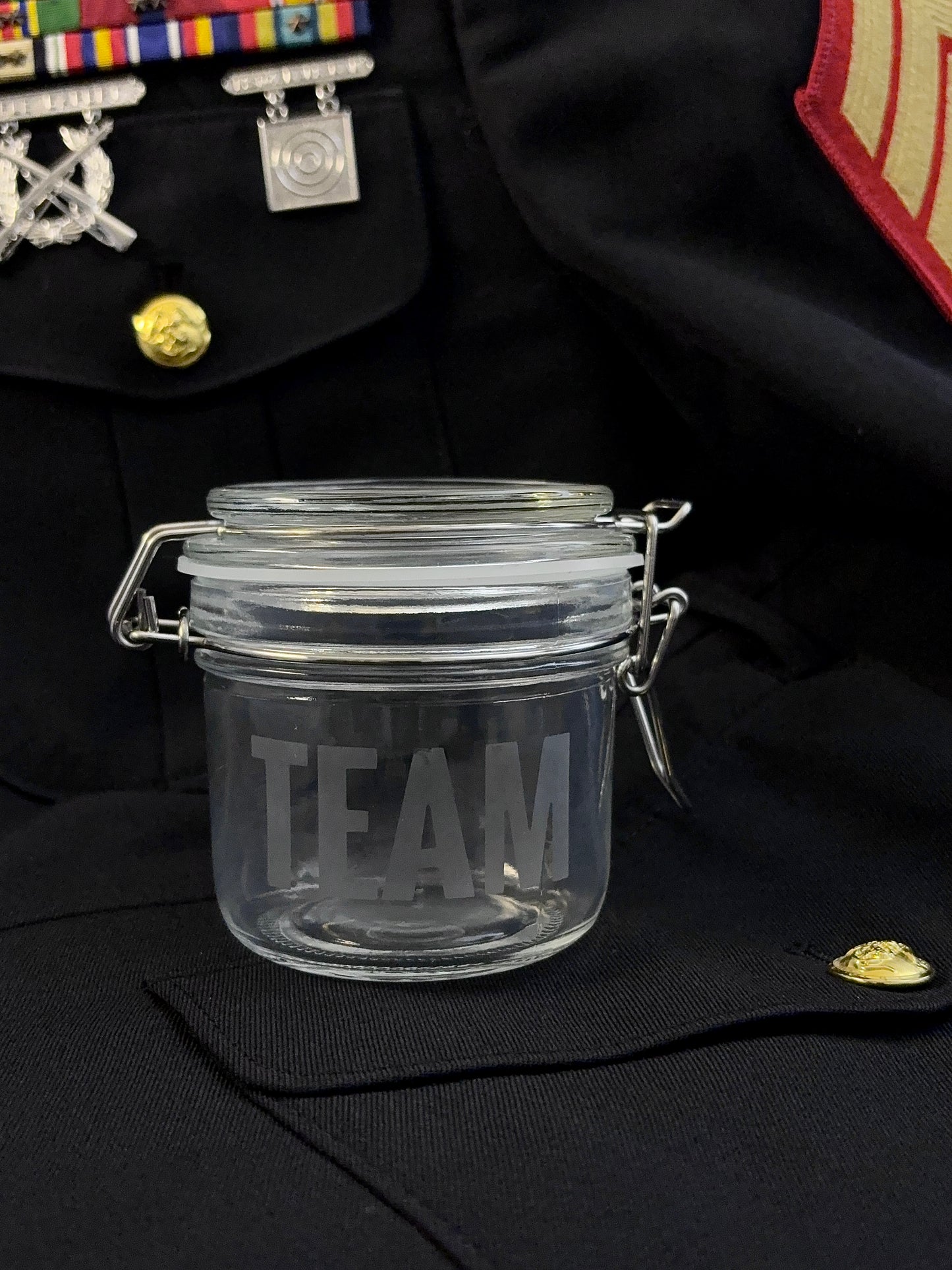 Clear glass jar with 'TEAM' etching on a black marine corps uniform background